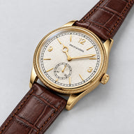 Specht & Söhne Invictus 1986 Gold em três quartos, com caixa dourada, mostrador branco, numerais dourados e pulseira marrom.